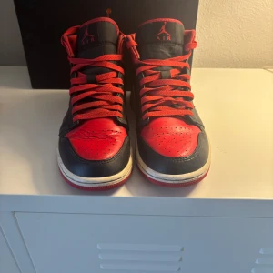 Nike Air Jordan 1 mid röd/svart - Säljer ett par Nike Air Jordan 1 High i klassisk röd och svart färg med vita detaljer på sulan. Skorna har röda snören och den ikoniska Jordan-loggan på plösen. Ovandelen är i skinn och sulan är röd med Nike-mönster undertill.
