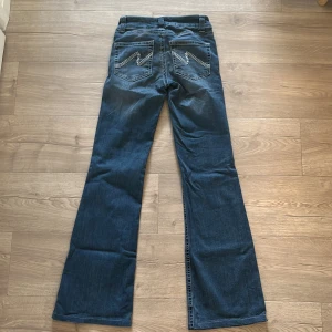 Trendiga blå bootcut jeans med broderade fickor - Snygga blå jeans med lågmidjad bootcut passform och snygga broderade detaljer på bakfickorna. Klassisk femficksmodell med dubbla knappar framtill. 