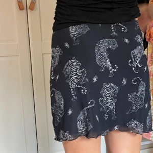 Svart kortkjol med tigerprint från H&M - Cool svart kortkjol från H&M Divided i storlek XS med ett unikt vitt tigerprint över hela kjolen. Kjolen har lätt vågad kant nedtill och är gjord i ett mjukt syntetmaterial som sitter skönt och har en normal passform. Perfekt för dig som gillar djurmotiv och vill sticka ut.