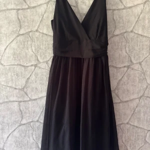 Svart långklänning från Vero Moda - Snygg svart långklänning från Vero Moda med omlott upptill och v-ringad hals. Klänningen har ett markerat midjeband och ett lätt, flytande tyg som ger en elegant siluett. Perfekt för dig som gillar stilrena och klassiska looks.