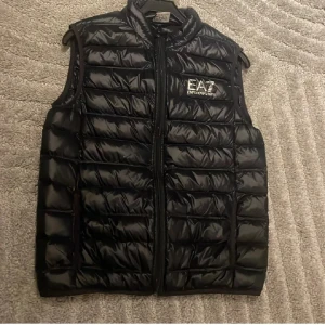 Svart dunväst från Emporio Armani EA7 - Snygg svart dunväst från Emporio Armani EA7 med quiltad design och dragkedja framtill. Västen har en stilren look med logga på bröstet och hög krage. Perfekt att slänga över en hoodie eller tröja för extra värme.