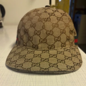 Gucci keps med GG-mönster och ränder - Snygg keps från Gucci i beige och brunt med klassiskt GG-monogram över hela kepsen. På sidorna finns gröna och röda ränder som ger den ikoniska Gucci-looken. Justerbar rem bak i brunt läder. Perfekt accessoar för att lyfta din outfit.