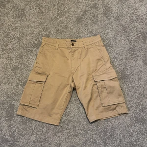 Beige cargo shorts från GANT W29 - Snygga beige cargo shorts från GANT med relaxed passform och flera praktiska fickor på sidorna och bak. Klassisk midja med knapp och dragkedja, tillverkade i mjuk bomull. Perfekta för chill dagar och sommarhäng.