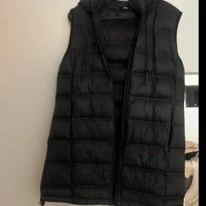 Svart dunväst från Zara - Svart dunväst från Zara med huva och dragsko. Västen har dragkedja framtill och är lätt vadderad för extra värme. Perfekt för lager-på-lager och har en clean, stilren look som funkar till det mesta.