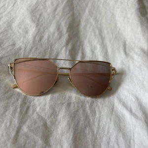 Guldiga solglasögon med rosa glas - Snygga solglasögon med guldiga metallbågar och spegelglas i rosa ton. Unik design med dubbla bågar upptill och tunna skalmar. Perfekta för dig som vill sticka ut med en trendig accessoar i sommar.