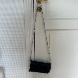 Svart axelväska med kedjerem - Snygg svart axelväska med silverfärgad kedjerem och kedjedetaljer längs kanterna. Väskan har en stilren, rektangulär form och är tillverkad i skinnimitation. Säljer mina väskor då de inte används, pris kan alltid diskuteras och tveka inte på att höra av dig om du har eventuella frågor💞