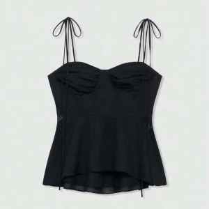 Svart korsett-topp med knytband - Supersnygg svart korsett-topp med tunna axelband som knyts i rosetter. Toppen har en lätt utsvängd nederdel och rynkade detaljer vid bysten för extra shape. Materialet är mjukt och känns lätt mot huden, perfekt för att skapa en cool och trendig look.