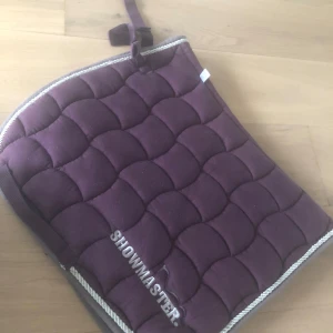 Quiltad vinröd schabrak från Showmaster - Quiltad schabrak i vinröd färg från Showmaster, passar till dressyrsadel för ponny. Snyggt mönstrad med ljus kant och broderad logga. Mjuk och stadig med praktiska fästen för sadelgjord.