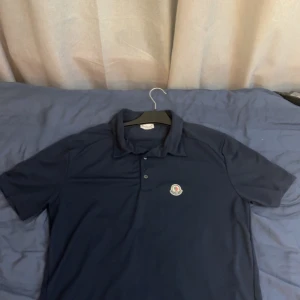 Marinblå pikétröja från Moncler - Snygg marinblå pikétröja från Moncler med klassisk krage och knäppning framtill. Tröjan har korta ärmar och Monclers ikoniska logga broderad på bröstet. Perfekt för en stilren och sportig look.