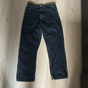 Svarta Carhartt Simple Pant 32x32 - Svarta byxor från Carhartt i modellen Simple Pant. De har raka ben, kontrastsömmar och klassisk Carhartt-logga på bakfickan. Tillverkade i bomull med lite polyester för extra slitstyrka. Perfekta för dig som gillar streetwear och workwear-vibe.