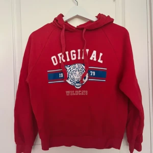 Röd hoodie Wildcats Gina Tricot XS - Snygg röd hoodie från Gina Tricot i storlek XS med tryck 'ORIGINAL WILDCATS' och en cool tiger på bröstet. Tröjan har huva med snörning och långa ärmar. Materialet är mjuk bomull och insidan är fleece. 