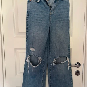 Blå ripped wide jeans från Gina Tricot - Säljer ett par ljusblå jeans från Gina Tricot med breda ben och stora slitningar över knäna. Modellen är high waist och har klassiska fem fickor. Perfekta för en avslappnad och trendig look.