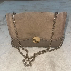 Beige mocka axelväska med kedja - Snygg beige axelväska i mocka och skinnimitation med silverfärgad kedjerem och rund metallknapp framtill. Stilren design med mjuk ovandel och slät botten, perfekt för att lyfta din outfit.