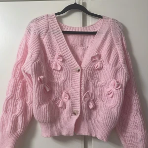 Rosa stickad kofta med rosetter - Supersöt ljusrosa stickad kofta med dekorativa rosetter framtill och pärlemorknappar. Koftan har ett unikt strukturmönster, ribbade muddar och lång ärm. Perfekt för dig som gillar gulliga detaljer och vill ha en mysig, trendig look. Den har fått någon liten fläck på armen men inget synbart, använd ett fåtal gånger.