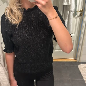 Glittrig svart stickad tröja med pärldetaljer - Supersnygg svart stickad tröja med glittriga inslag och korta puffärmar. Tröjan har en liten volang vid halsen och pärlformade knappar på ena axeln som extra detalj.
