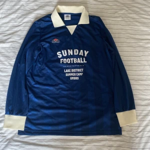 Blå långärmad fotbollströja Umbro StM - Snygg blå långärmad fotbollströja från Umbro med vit krage och muddar. Tryck på bröstet med texten 'Sunday Football' och 'Lake District Summer Camp'. Tillverkad i 100% polyester. Klassisk retrostil med diskreta ränder i tyget och i mkt bra skick. 