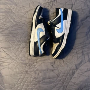 Nike Dunk Low svart/vit/blå strl 44 - Nike Dunk Low sneakers i svart, vitt och ljusblått. Klassisk siluett med perforerad tå, svart snörning och kontrasterande blå swoosh. Skorna har platt sula och är tillverkade i skinn och syntet. Perfekt för dig som gillar streetstyle och vill sticka ut.