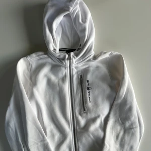 Vit hoodie från Sail Racing med dragkedja - Vit hoodie från Sail Racing med hel dragkedja och justerbar huva. Tröjan har en bröstficka med dragkedja och Sail Racing-logga på bröstet. Perfekt för en clean och sportig look!