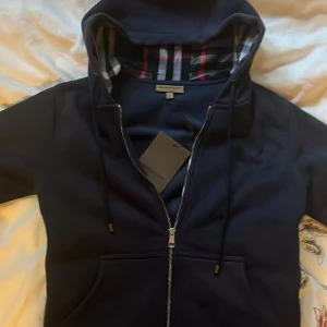 Marinblå huvtröja från Burberry - Säljer en stilren marinblå huvtröja från Burberry med klassiskt rutigt mönster i huvan. Tröjan har dragkedja framtill och en broderad logga på bröstet. Perfekt för en casual look med en touch av lyx. Mycket bra skick!🧥✨