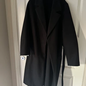 Svart kappa från Vero Moda, storlek L - Stilren svart kappa från Vero Moda i storlek L. Klassisk design med krage, knytband i midjan och diskreta fickor. Tillverkad i mjuk polyester som känns skön och passar perfekt till höst och vinter. Enkel att matcha med allt i garderoben.