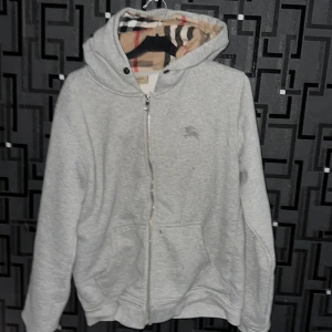Grå Burberry hoodie med dragkedja - Säljer en grå hoodie från Burberry i storlek M. Tröjan har klassisk dragkedja, huva med det ikoniska rutiga Burberry-mönstret på insidan och två fickor framtill. Perfekt för dig som gillar stilren design med lyxig touch och vill ha något som sticker ut.