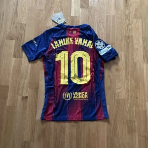 FC Barcelona Lamine Yamal #10 matchtröja (SPELARVERSION) - Säljer en officiell FC Barcelona fotbollströja med Lamine Yamal och nummer 10 på ryggen. Tröjan har klassiska blå och röda ränder, gult tryck, Champions League-märke på ärmen och sponsorloggor från Spotify och UNHCR. Tillverkad i lätt polyester med korta ärmar och atletisk passform. (PLAYER EDITION)