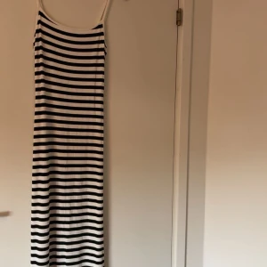 Randig långklänning med smala axelband - Säljer en långklänning med svarta och vita horisontella ränder. Klänningen har smala axelband och är figurnära med en rak siluett. Materialet är ribbat och stretchigt, perfekt för varma dagar. Enkel och stilren design som känns fräsch och modern. Aldrig använd. 