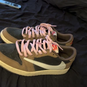 Nike Jordan 1 Low Travis Scott - Nike Jordan 1 Low Travis Scott med bruna och svarta paneler i mocka och skinn, vit omvänd swoosh och rosa snören. Röd broderad logga på hälen och klassisk Air Jordan-vinge. Snygg och unik färgkombo som sticker ut.