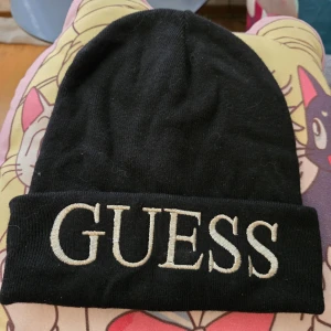 Svart mössa från Guess - Svart ribbstickad mössa från Guess med bred uppvikt kant och stor vit broderad logga framtill. 