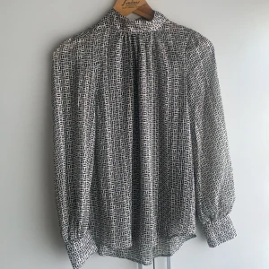 Vit och svart mönstrad blus från H&M - Snygg blus från H&M i storlek S med svart abstrakt mönster på vit botten. Blusen har hög krage, knappar i nacken och långa ärmar med breda manschetter. Materialet är lätt och luftigt, perfekt för att ge en cool vibe till din outfit.