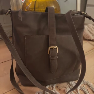 Brun axelväska med blommigt foder - Snygg brun axelväska i mocka med justerbar rem och guldfärgat spänne framtill. Väskan har en praktisk ficka på framsidan och dragkedja på baksidan. Insidan är fodrad med blommigt tyg i färger som orange, blå och röd. Står inte vart den är ifrån! 