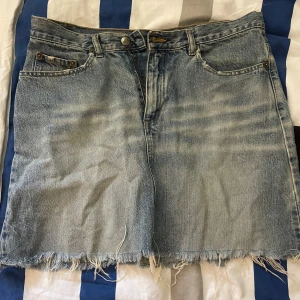 Blå jeanskjol med rå kant från Divided - Säljer en ljusblå jeanskjol från Divided i storlek 38. Kjolen har klassisk femficksdesign, dragkedja och knapp framtill samt en rå, fransig kant nedtill för en cool vibe. Perfekt att matcha med sneakers eller boots.