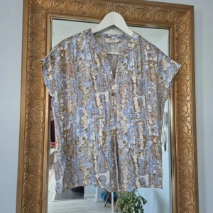 Blå och beige mönstrad skjorta Cream - Kortärmad skjorta från Cream i storlek 36 med ett unikt mönster i blått, beige och vitt. Skjortan har v-ringad hals och knappar framtill. Materialet är mjukt och lätt, perfekt för varmare dagar. Snygg och avslappnad passform som ger en cool vibe.