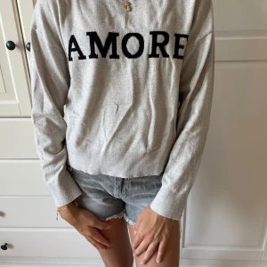 Beige tröja med AMORE från Gina Tricot - Säljer en beige långärmad tröja från Gina Tricot med svart text 'AMORE' framtill. Tröjan har en rund hals och är tillverkad i mjuk bomullsmix. Perfekt för dig som gillar enkel stil med en cool detalj framtill.