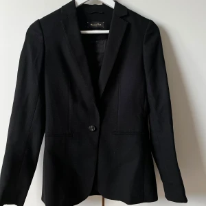 Svart kavaj från Massimo Dutti - Stilren svart kavaj från Massimo Dutti med italienskt tyg, tillverkad i Portugal. Klassisk modell med en knapp framtill, smal krage och diskreta fickor. Passformen är normal och materialet känns som ullblandning. Perfekt för dig som gillar en clean och elegant look.