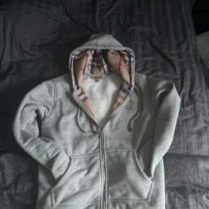 Grå hoodie från Burberry med rutig huva - Säljer en grå hoodie från Burberry med klassisk rutig insida i huvan och broderad logga på bröstet. Tröjan har dragkedja, snörning i huvan och två fickor framtill. Perfekt för dig som gillar stilrena och lyxiga streetwear-plagg.