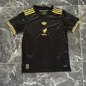 Svart Mexiko fotbollströja Adidas - Snygg svart Mexiko fotbollströja från Adidas med guldiga detaljer och tryck. Tröjan har korta ärmar med gulddetaljer och tre guldiga ränder på axlarna. Materialet är lätt och andas, perfekt för träning eller match. Aeroready-teknologi för extra komfort.