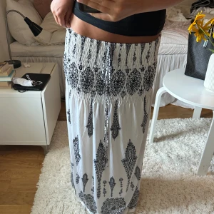Vit långkjol med svart mönster - Säljer en vit långkjol med svart paisley-inspirerat mönster. Kjolen har smock i midjan och snörning framtill för en skön passform. Materialet känns lätt och luftigt, perfekt för varma dagar. Mönstret ger en cool boho-vibe och kjolen är riktigt flowy.