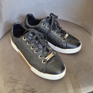 Svarta sneakers Tommy Hilfiger - Snygga svarta sneakers från Tommy Hilfiger med vit sula och gulddetaljer. Sidorna har ett coolt geometriskt mönster och metallplatta med logga. Skorna har snörning och rund tå, samt en liten logga bak på hälen. Perfekta för dig som gillar stilrena och trendiga skor.