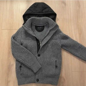 Grå stickad cardigan jacka Marc O'Polo - Snygg grå stickad cardigan jacka från Marc O'Polo med grov ribbstickning, svarta knappar och dragkedja. Jackan har en svart avtagbar huva i syntetmaterial och två fickor framtill. Perfekt för kyliga dagar och riktigt skön att bära.