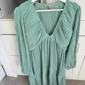 Ljusgrön plisserad klänning från H&M - Säljer en ljusgrön plisserad klänning från H&M med v-ringning och långa ärmar. Klänningen har en flowy passform och snygga volangdetaljer nedtill. 