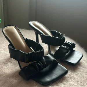 Svarta klackade sandaler med rynkade band - Säljer ett par svarta sandaler med hög klack och rynkade band över foten. De har en fyrkantig tå och är tillverkade i mjukt skinnliknande material. Perfekt för dig som gillar statement-skor med cool design och vill sticka ut lite extra.
