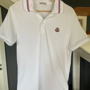 Vit Moncler piké med röd/blå krage - Vit Moncler t-shirt i pikémodell med korta ärmar och klassisk krage med röd och blå rand. Broderad Moncler-logga på bröstet och knappar vid halsen. Tillverkad i mjuk bomull som känns skön mot huden. Perfekt för dig som gillar stilrena och sportiga plagg. Den är köpt i Göteborg NK men tyvärr tappat bort kvittot. Ny pris: 3599 knappt använt den