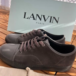 Lanvins cap toe - Snygga grå sneakers från Lanvin med ovandel i mjuk mocka och tåparti i slät skinn. Skorna har rund tå, platt sula och klassisk snörning. Perfekt för dig som gillar stilrena och exklusiva sneakers med en lyxig känsla. Dustbag och originalkartong medföljer.
