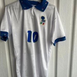 Italien R. Baggio VM 94 fotbollströja - Säljer en ikonisk vit fotbollströja från Italiens landslag med blå detaljer, nummer 10 och R. Baggio på ryggen. Tröjan har korta ärmar, blå krage och muddar med färgglada trianglar. Materialet är lätt polyester, perfekt för match eller samling.