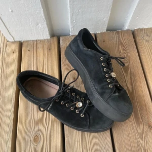 Svarta mockasneakers med platt sula - Snygga svarta sneakers från Dasia i mjuk mocka med platt sula och rund tå. Skorna har guldfärgade öljetter och snörning med en dekorativ pärldetalj på snöret. Perfekta för dig som gillar stilrena och coola skor med lite extra bling.