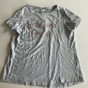 Grå t-shirt från lager 157 - Grå t-shirt med rund hals och korta ärmar. Framsidan har ett rosa tryck med texten 'Wild and free' och en fjäder. T-shirten är i mjuk bomull och har en avslappnad passform, perfekt för chill dagar.