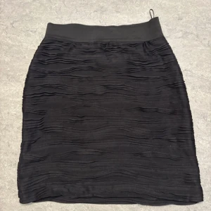 Svart kortkjol från H&M med struktur - Svart kortkjol från H&M i storlek 38. Kjolen har ett coolt strukturerat tyg med vågiga detaljer som ger en unik look. Den har bred resår i midjan. Perfekt för dig som gillar stilrena plagg.