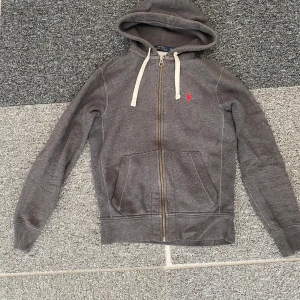 Grå hoodie från Polo Ralph Lauren XS - Säljer en grå hoodie från Polo Ralph Lauren i storlek XS. Tröjan har dragkedja, huva med vita snören och ett rött Polo-logo broderat på bröstet. Mjuk bomullskänsla och praktiska fickor framtill. Perfekt för chill dagar och streetwear vibes.