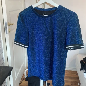 Blå glittrig t-shirt från Vero Moda - Snygg blå t-shirt från Vero Moda med glittrigt tyg och svarta samt vita ränder på ärmsluten. Modellen är klassisk med rund hals och korta ärmar, perfekt för dig som gillar att sticka ut med lite extra shimmer. Materialet känns mjukt och har en cool glansig finish.